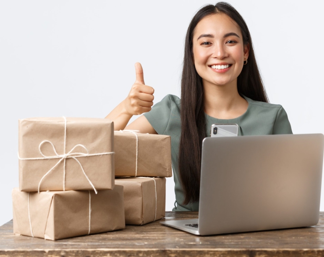 Happy online business owner, with online shop, packing orders, show thumbs-up, manage clients requests via laptop.